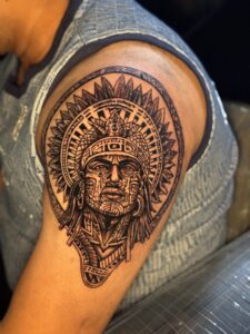 Black and grey tattoo of an Aztec warrior head with feathered headdress on shoulder — 1920 Tattooz Hub, Ahmedabad