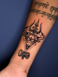 Black and grey tattoo of a bull skull with trishul and tabla drum on forearm — 1920 Tattooz Hub, Ahmedabad