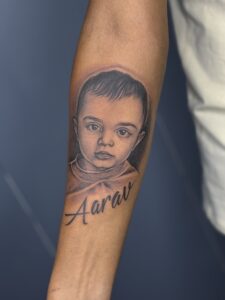 Black and grey tattoo of a child's portrait with name lettering on forearm — 1920 Tattooz Hub, Ahmedabad