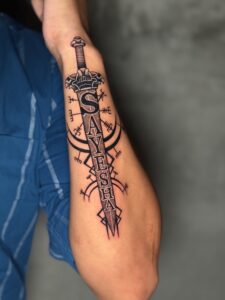 Black and grey tattoo of a dagger with name lettering and a compass symbol on forearm — 1920 Tattooz Hub, Ahmedabad