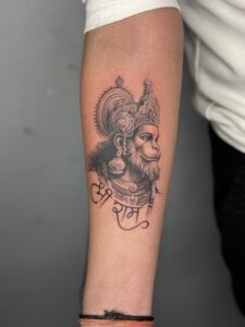 Black and grey tattoo of Hanuman with an orange tilak accent on forearm — 1920 Tattooz Hub, Ahmedabad
