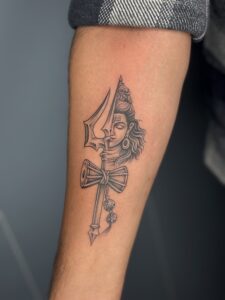 Black and grey tattoo of a trishul merged with Lord Shiva's face and a damru on forearm — 1920 Tattooz Hub, Ahmedabad