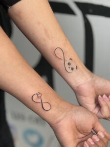 Fine line tattoo of matching infinity symbols with a panda and a bow on wrist — 1920 Tattooz Hub, Ahmedabad