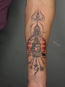 Realistic tattoo of Lord Shiva with trishul, damru, and a sun backdrop on forearm — 1920 Tattooz Hub, Ahmedabad